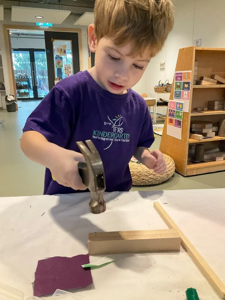 A young boy using a hammer on a small wooden piece in a classroom or activity space.