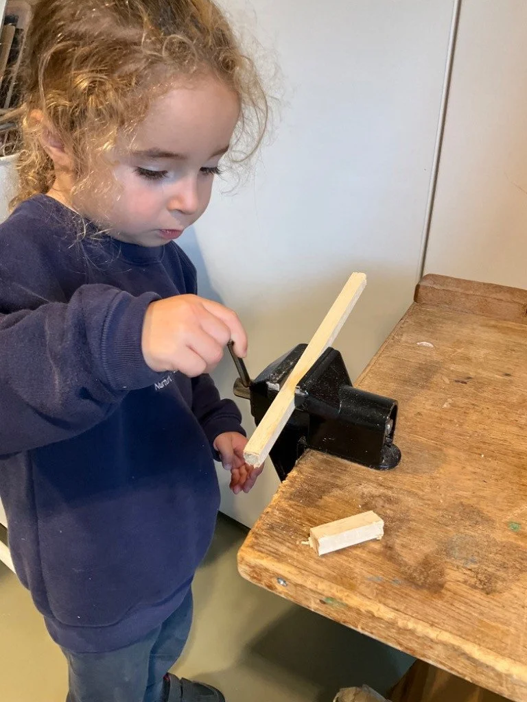 Young girl operating a woodworking vise on a wooden workbench, holding a piece of wood with a tool, in an indoor setting.