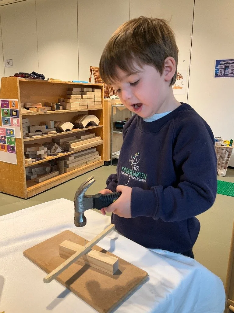 A young boy with brown hair wearing a navy blue sweatshirt, holding a small hammer, and working on a woodworking project with wooden blocks and a stick. woodwork for math development. EYFS 
