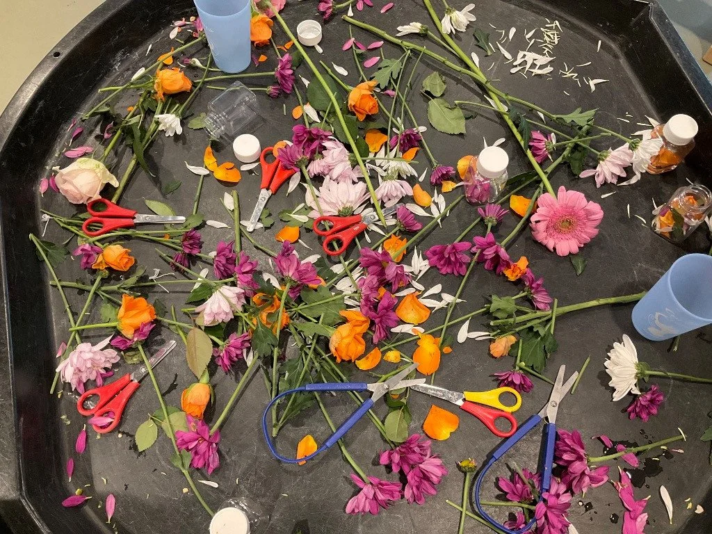 A messy black table with various flower stems, leaves, petals, scissors, and plastic cups. Some loose flower petals are scattered across the surface.