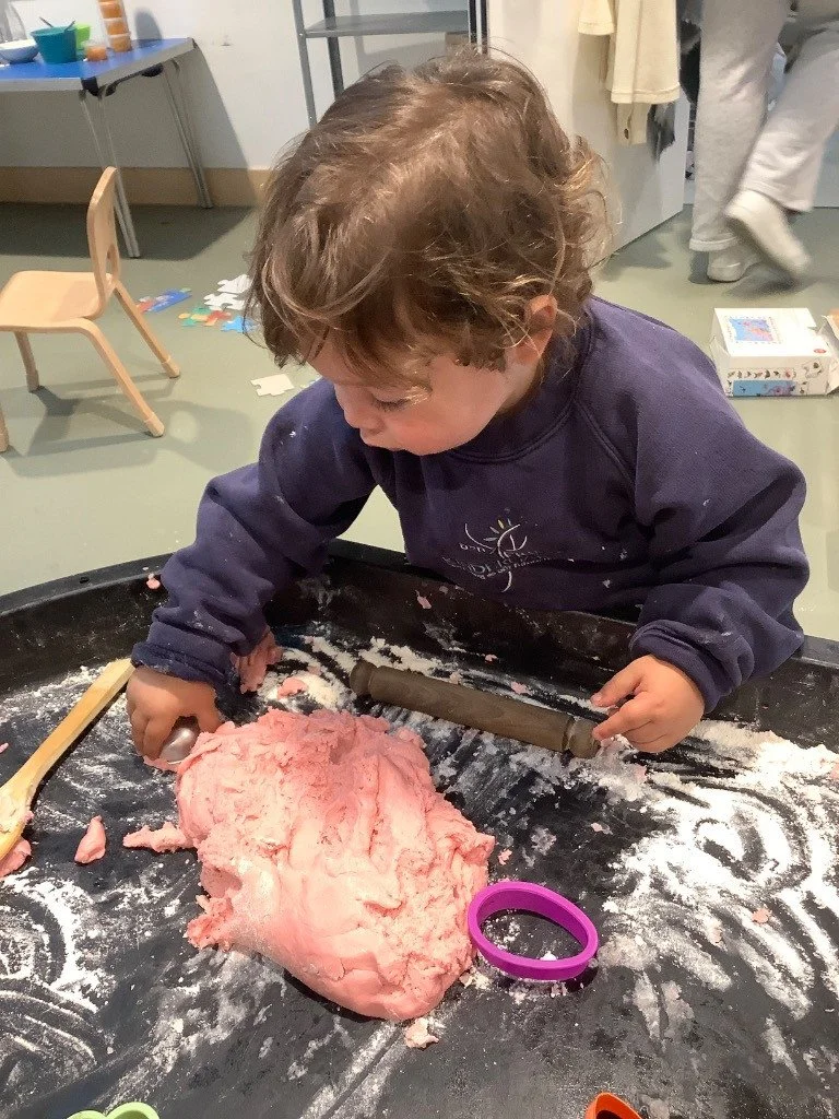 A young child with curly hair wearing a navy sweatshirt is playing with pink playdough on a black table. The playdough is shaped into a large mound, and the child is using a rolling pin to flatten it. There are some toys, a chair, and other children 