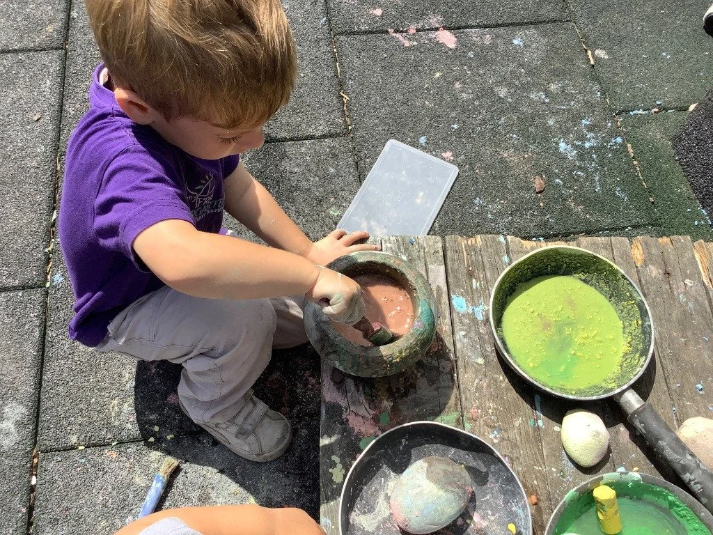 A young boy is sitting outdoors on a pavement, working on a craft project involving a bowling ball and paint. There are bowls with colorful paint and a small container nearby.