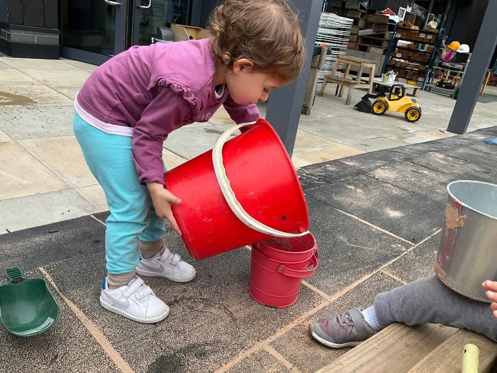 A young girl is pouring dirt from a large red bucket into a smaller pink bucket. She is outdoors on a tiled surface, wearing a purple jacket, light blue leggings, and white sneakers. There are other buckets and toys nearby, with some building materia