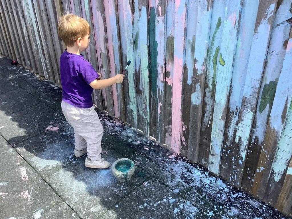 Young boy with a paintbrush standing on sidewalk, painting on a wooden fence with light pastel colors, with paint splatters on the ground.
