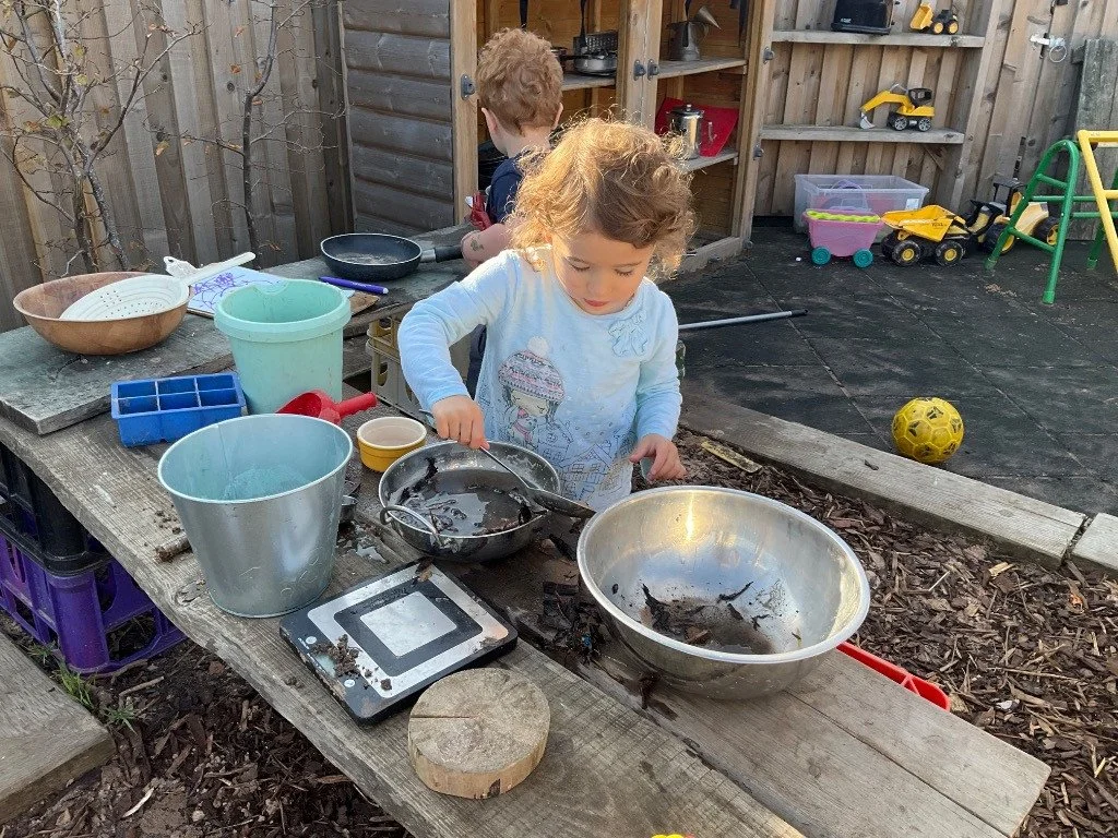 Two young children playing with water and mud in an outdoor wooden play area, surrounded by various toys and containers.