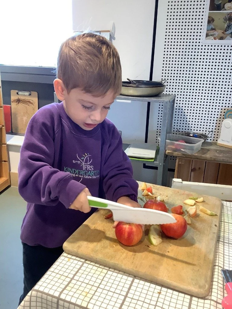 A young boy chopping apples on a wooden cutting board in a kitchen or classroom setting.