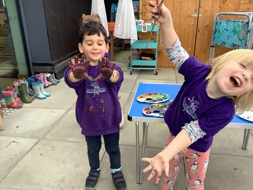 Two young children in purple shirts with the kindergarten logo, showing off their painted hands and smiling. One child has hand-painted hands, and the other is holding a paintbrush with paint on her hand, in a classroom setting with shoes lined up in the background.