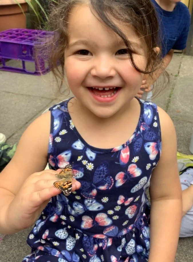 A young girl with a big smile holding a butterfly on her finger, outdoors with a purple crate in the background.