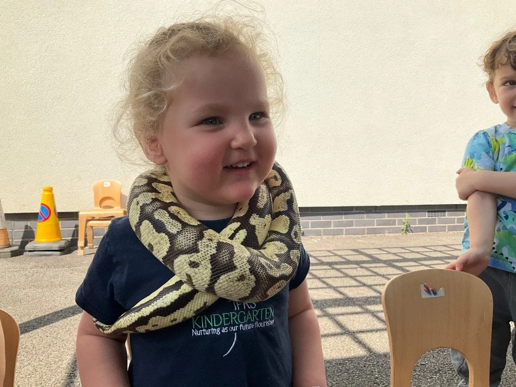 A young girl with curly blonde hair smiling as a python snake is wrapped around her neck outdoors on a sunny day. Two other children are partially visible nearby.