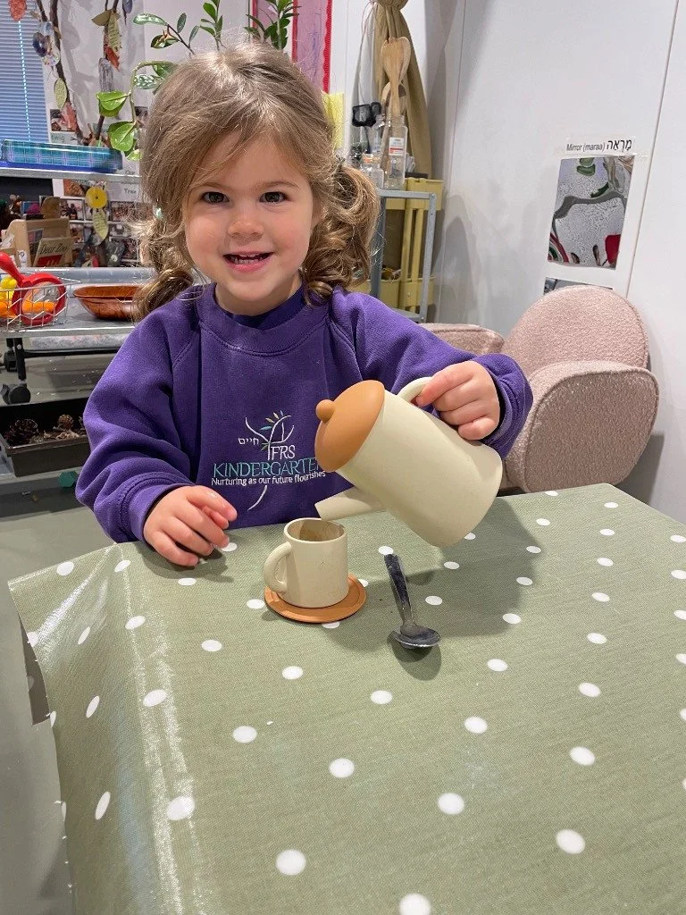 A young girl with curly hair pouring a liquid from a bear-shaped teapot into a mug on a polka dot tablecloth. Role play in a classroom, nursery in finchley london.