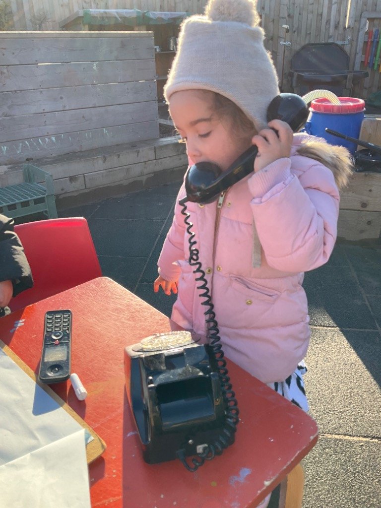 A young girl in a pink jacket and white knit hat using a black rotary phone on an outdoor table.