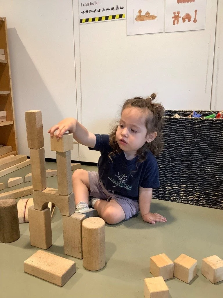 A young girl with curly hair, wearing a navy T-shirt and shorts, sits on the floor playing with large wooden building blocks, stacking them into a tower.