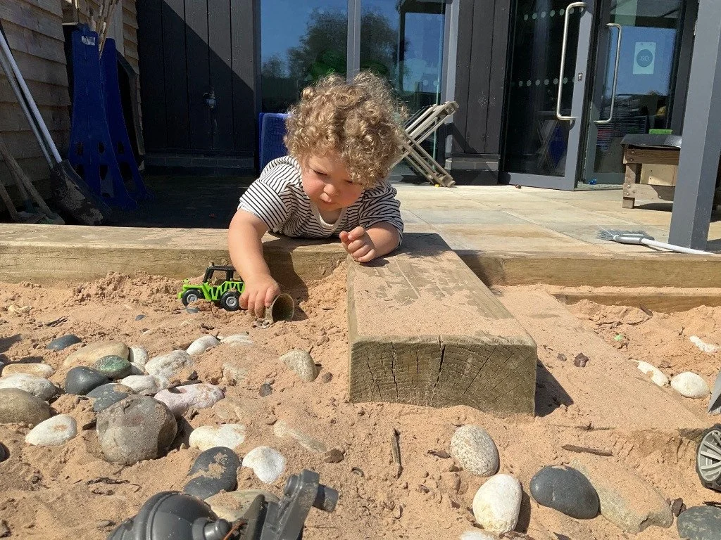 A young child with curly hair playing in a sandbox outside a building, surrounded by rocks and toys.