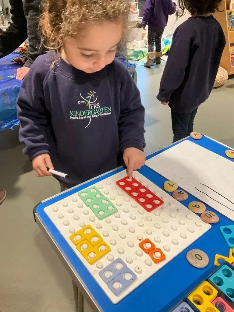 A young girl with curly hair wearing a preschool sweatshirt is playing with colorful letter tiles on a pegboard at a classroom table.