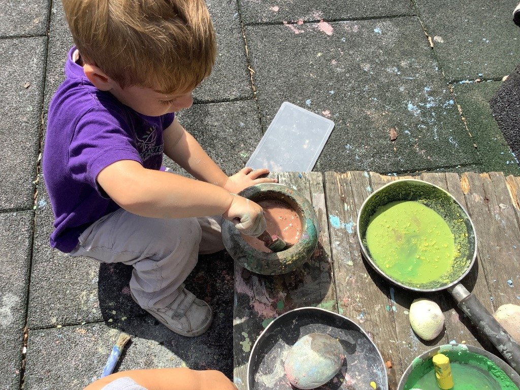A young boy wearing a purple shirt and beige pants is sitting on a outdoor pavement, using a tool to paint or work with colors inside a round container. There are also pans with bright green and dark colors, a white egg, and a yellow chalk or crayon on a wooden surface nearby.
