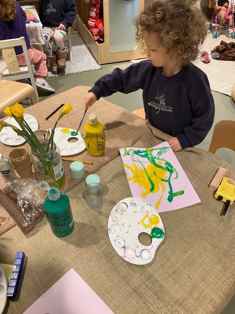 A young child with curly hair painting with green and yellow paints on a sheet of paper at a table. The table has painting supplies, including a palette, a bottle of yellow paint, and cups of water, with other children in the background participating in activities.