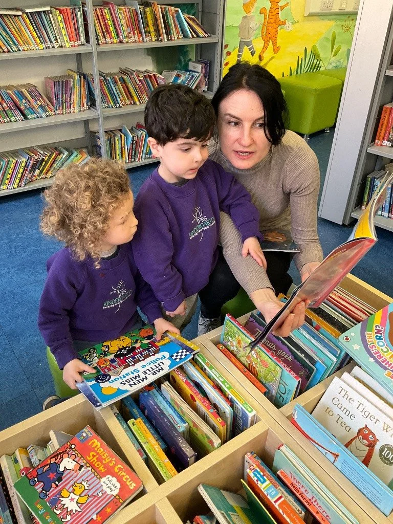 A woman and two children looking at a book together in a library, surrounded by shelves of children's books.