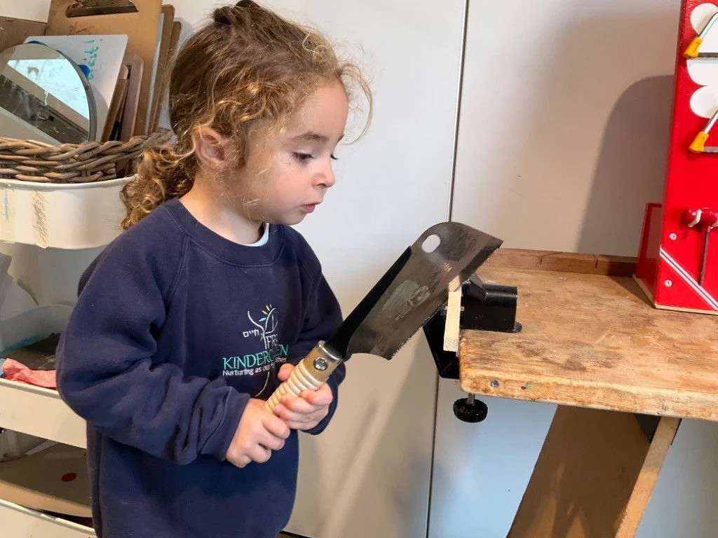 A young girl with curly hair is using a large metal spatula to press or flip something on a wooden workbench in a classroom or workshop setting.