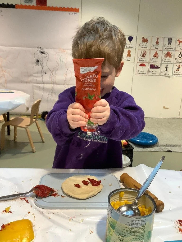 A young boy in a purple sweatshirt holding a tube of tomato purée in front of his face, preparing to make a pizza with pizza dough, tomato paste, canned corn, and rolling pin on a table in a classroom.