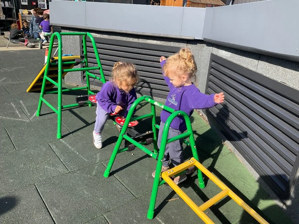 Two young girls in purple jackets playing on outdoor climbing equipment at a playground. Other children are in the background.