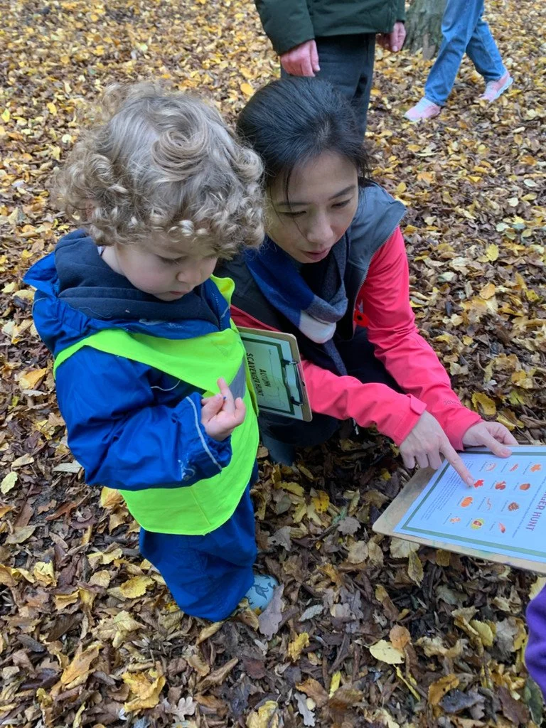 A woman and a young boy outdoors on a leaf-covered ground, looking at a learning sheet about nature or animals, with the woman pointing at the sheet.