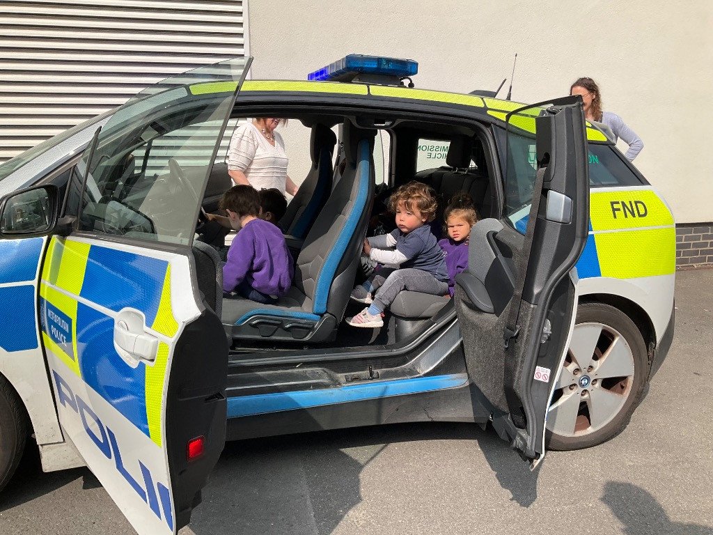 Children sitting inside a police car with open doors, with some adults standing nearby.