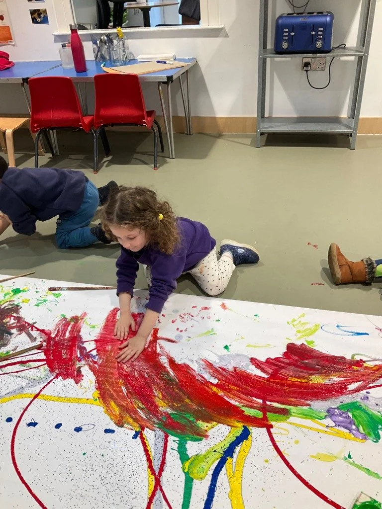 Young girl creating colorful abstract painting on large sheet of paper, lying on her stomach and pressing her hands into the paint, in a room with tables, red chairs, and a metal shelf.