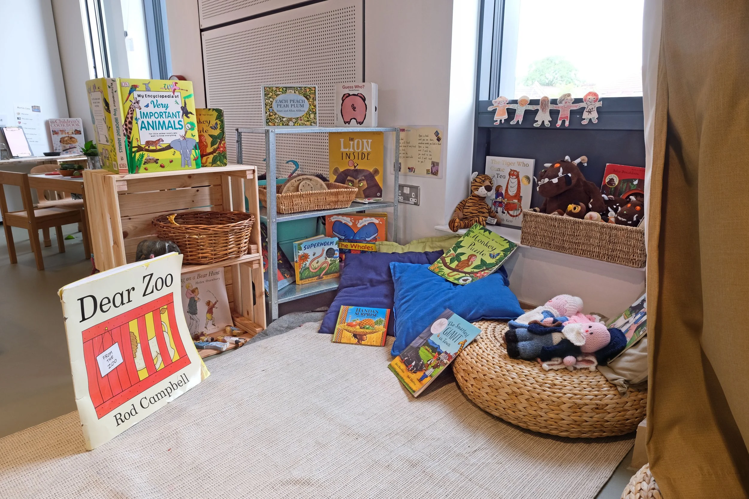Children's corner with books, stuffed animals, and cushions on a beige rug. A small wooden shelf holds children's books like "Dear Zoo" and "The Snail and the Giant". A basket and a small bookshelf display more books and plush toys, including a tiger and a bear. Window ledge has plush toys and more children's books. A string of paper doll decorations hangs above the window.