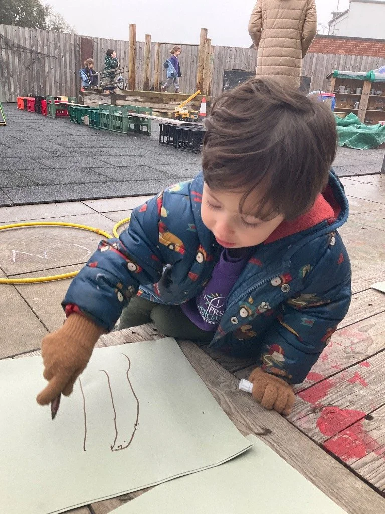Young boy drawing wavy lines on paper with a marker outdoors, others in background, wooden fence and play area.
