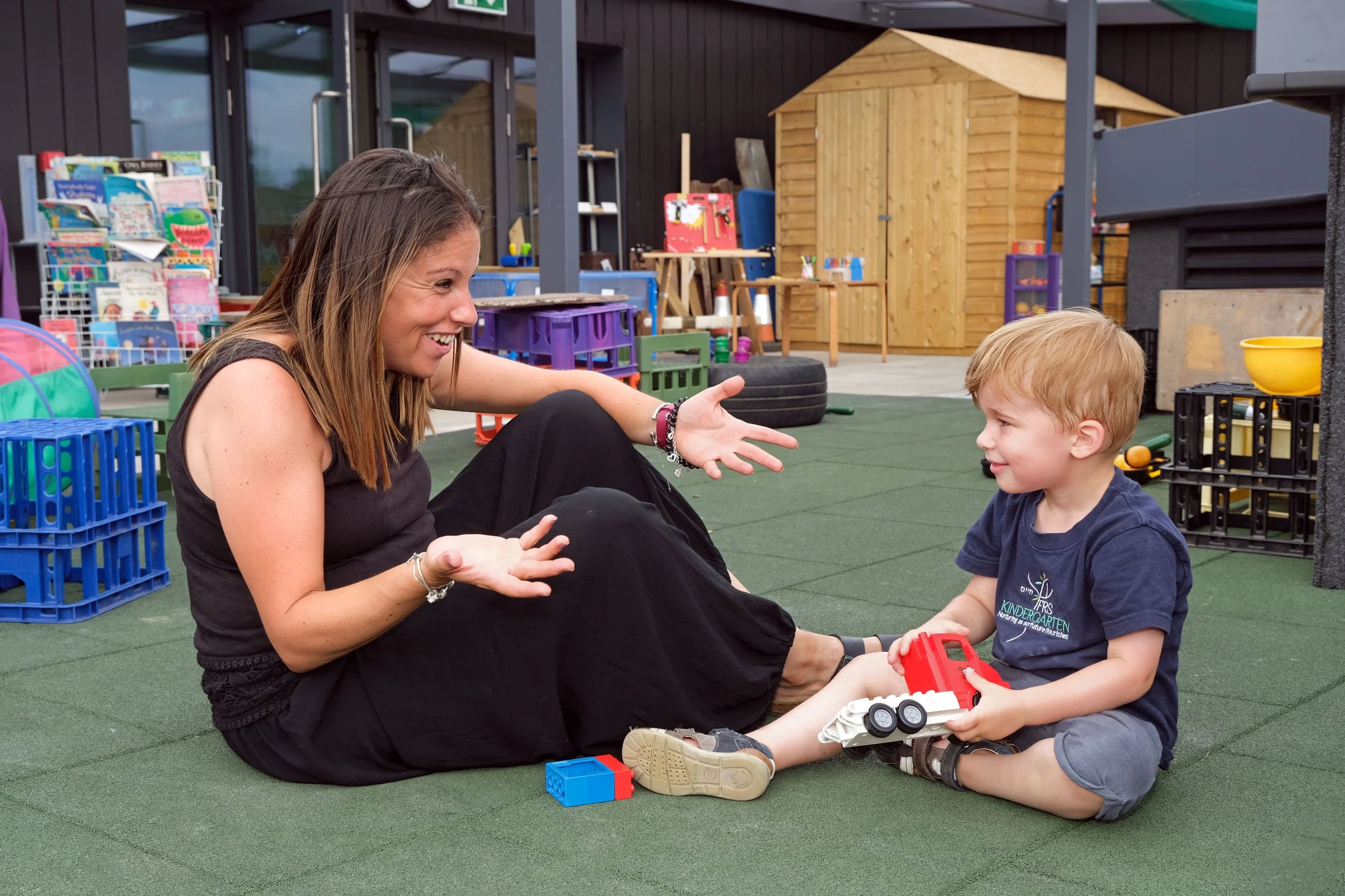 A woman and a young boy sitting on a playground mat, playing with toy trucks and enjoying a conversation.