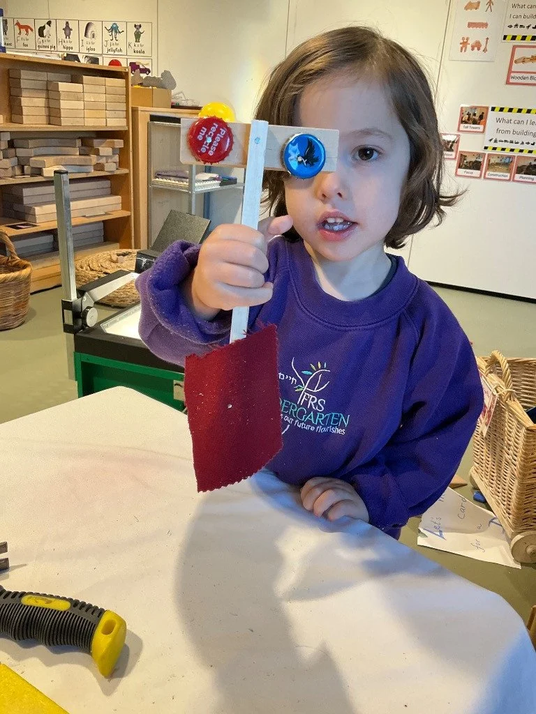 A young girl with shoulder-length brown hair wearing a purple sweatshirt is holding a homemade puppet made of a wooden stick, a felt piece, and two bottle caps with pictures of a fish and a blue whale. She is standing at a table in a classroom with e