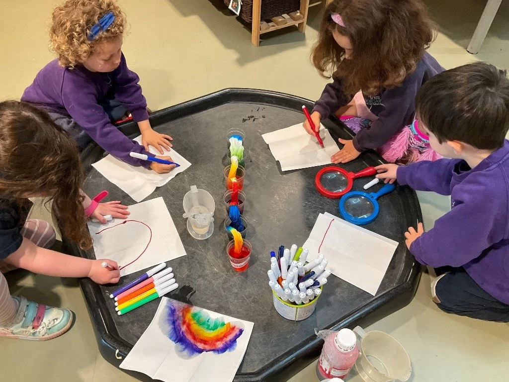 Five young children, four girls and one boy, are gathered around a black, circular table engaged in drawing and coloring activities with markers on white paper. The table has various art supplies, including markers, magnifying glasses, and cups of co