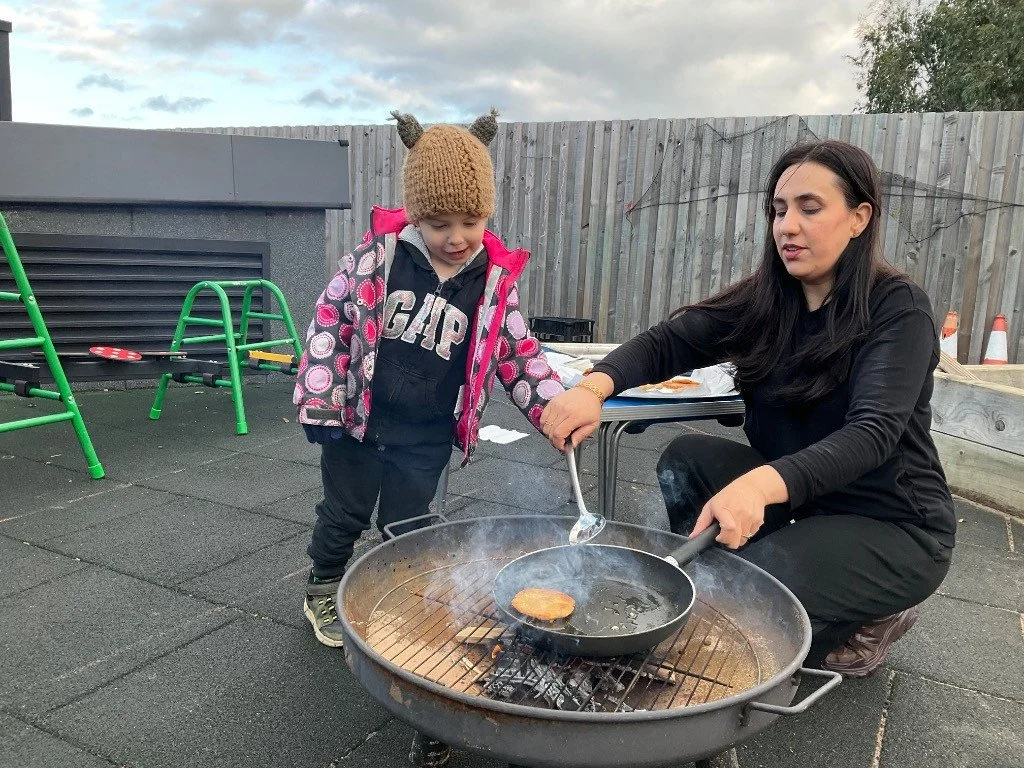 A woman and a young child outdoors, with the woman cooking a burger on a grill. The child is watching, wearing a brown knit hat with small ears and a colorful jacket. Outdoor play, child led experience.