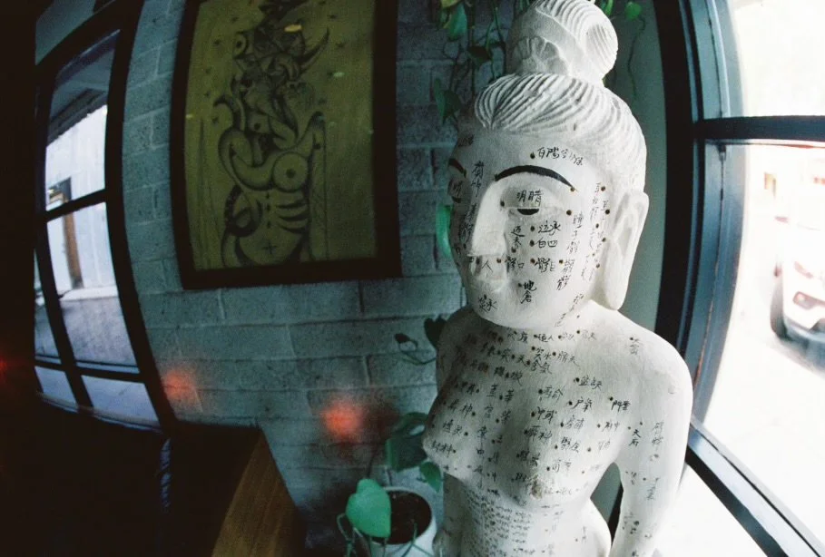 A white stone Buddha statue with writing on its face and body, placed within Dame of the West's lobby is next to a large window with a brick wall background and framed traditional tattoo flash designs.