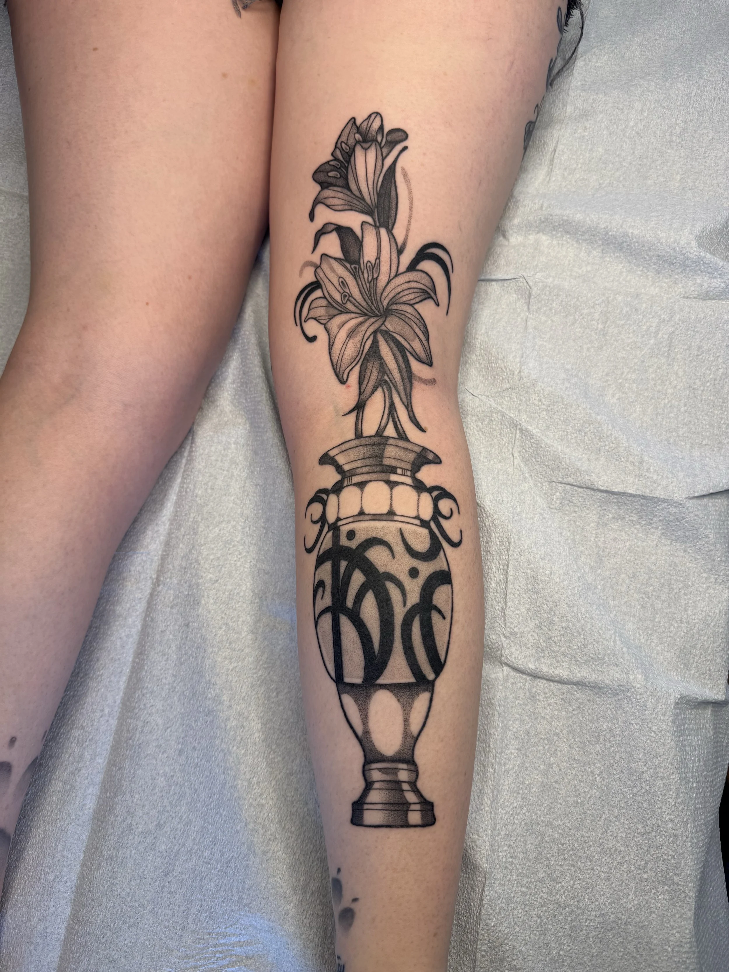 Tattoo artist Mel Gonzales, @theemelodic black and gray tattoo of lilies in a vase on the leg at Dame of the West Tattoo in Scottsdale, Arizona.