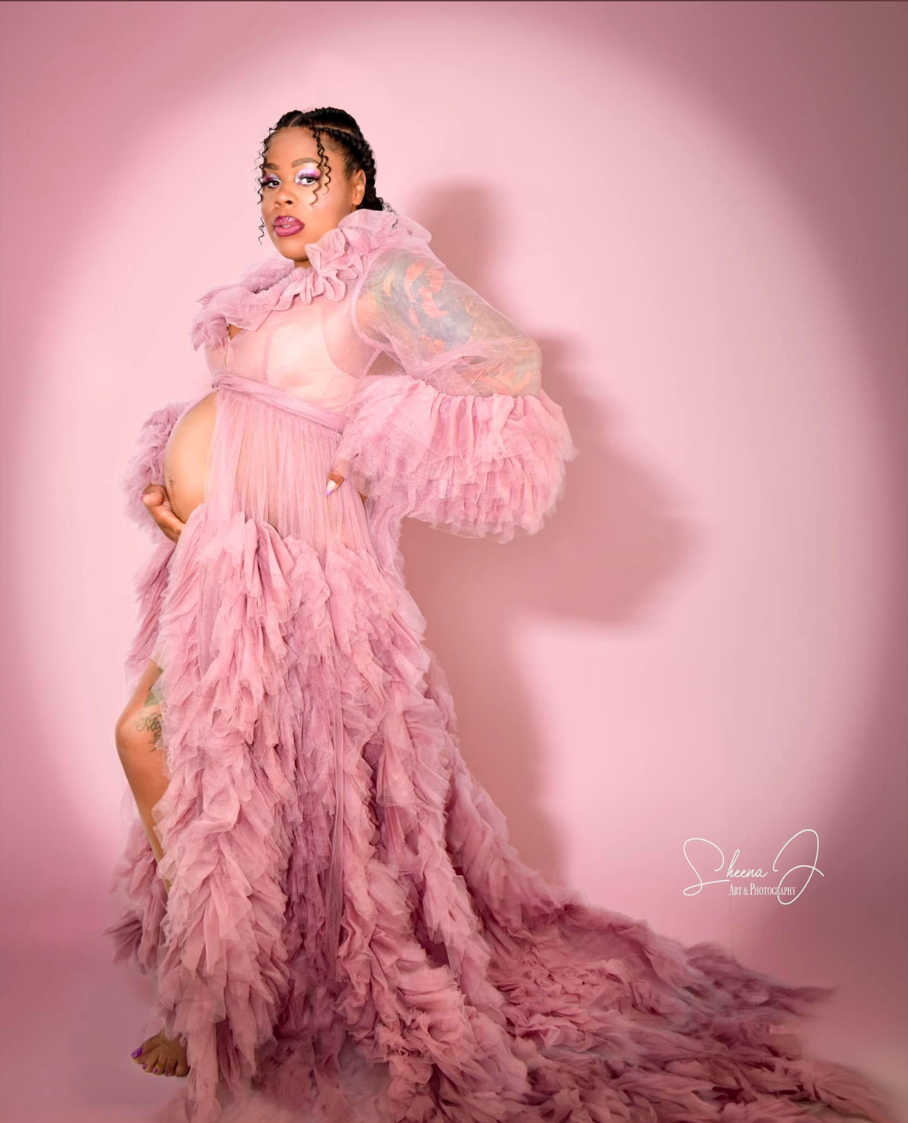 Pregnant woman in a pink, ruffled, flowing dress with sheer and lace details posing against a pink background.