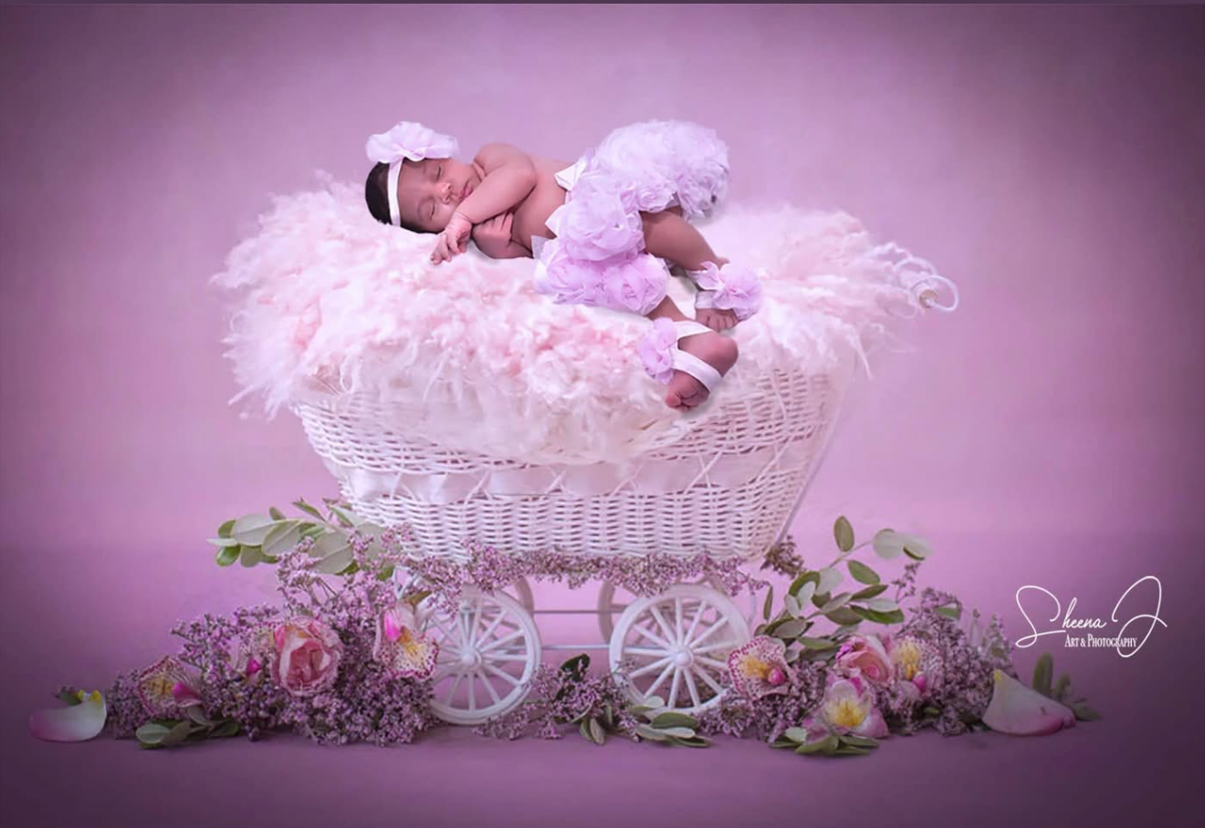 A newborn baby sleeping in a white wicker stroller decorated with pink and purple flowers and surrounded by pink and purple flowers, against a pink background.