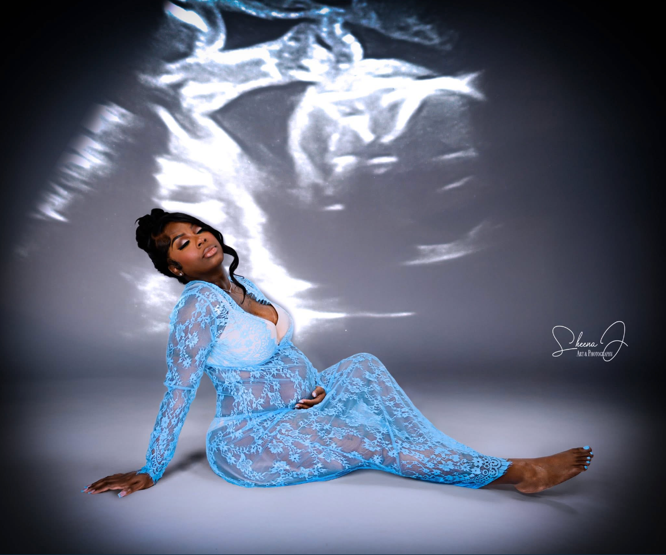 A woman in a blue lace dress sitting barefoot on the floor with her eyes closed, one hand on her pregnant belly, against a backdrop of a projected ultrasound image of a fetus.