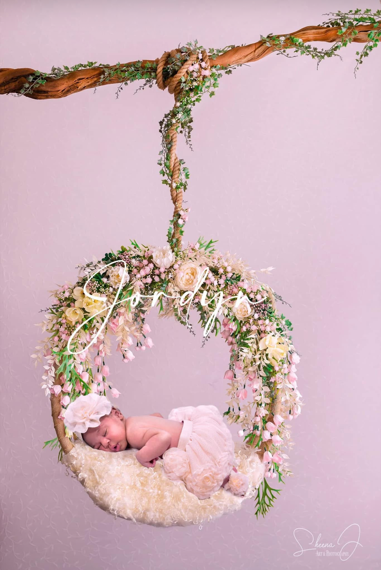 A sleeping baby girl on a floral swing decorated with pink and white flowers, hanging from a wooden branch with green vines, against a soft pink wall background.