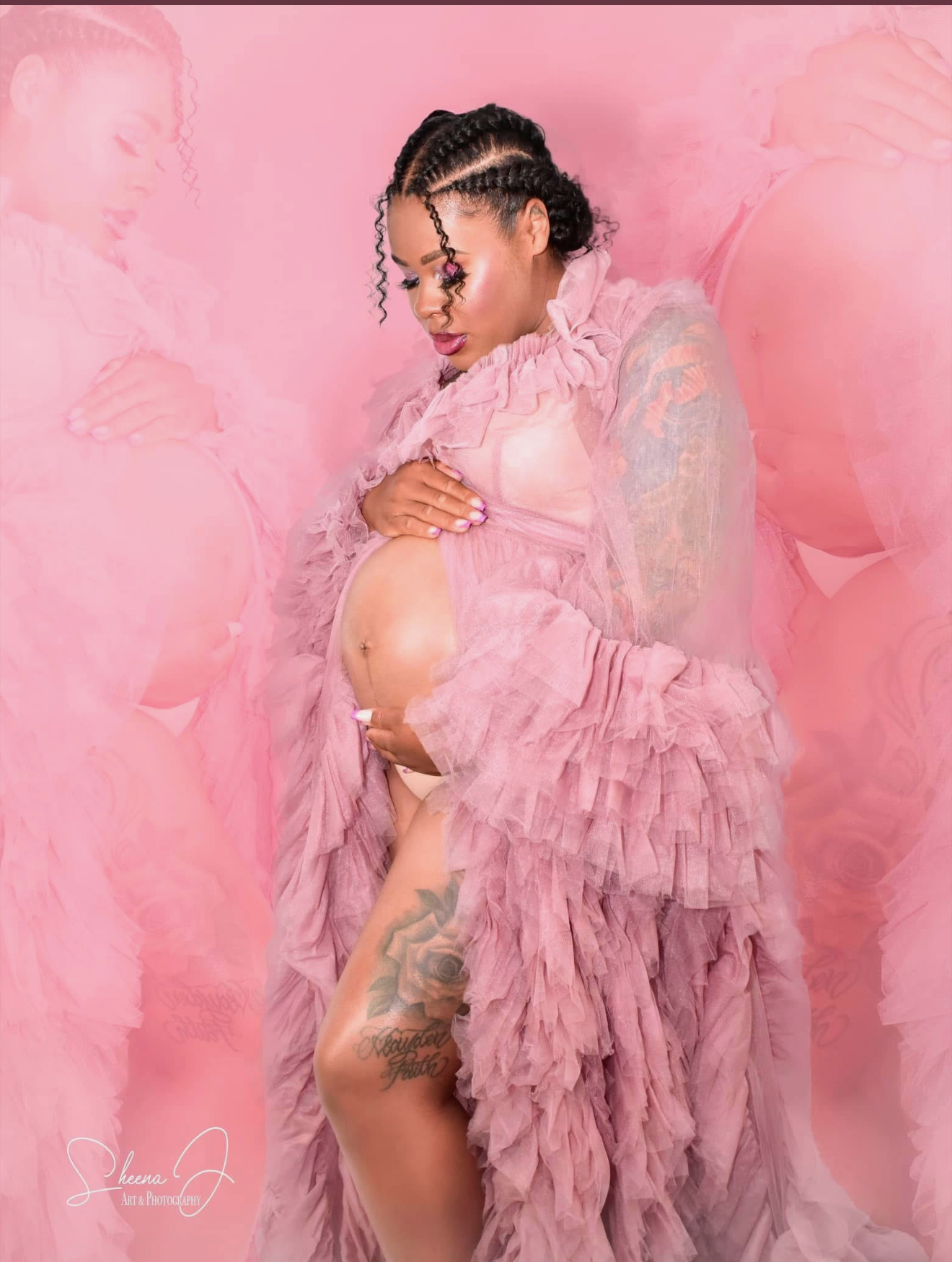 A pregnant woman in a pink ruffled dress is posed against a pink background, with her hand resting on her belly and the other hand on her thigh. She has dark curly hair styled in braids and is looking down at her belly.