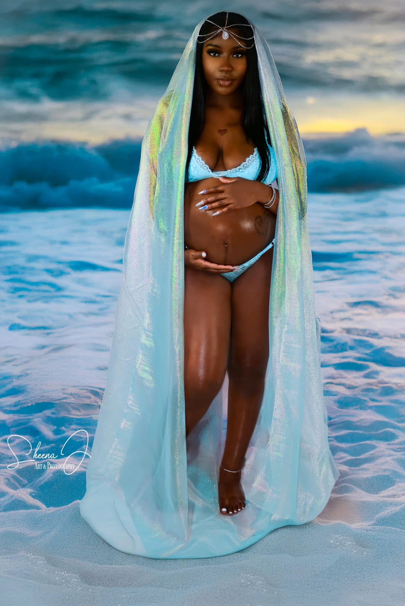 A woman standing on the beach with ocean waves in the background, wearing a blue bikini and a sheer, iridescent veil. She has long black hair, jewelry including a headpiece, and is touching her pregnant belly.