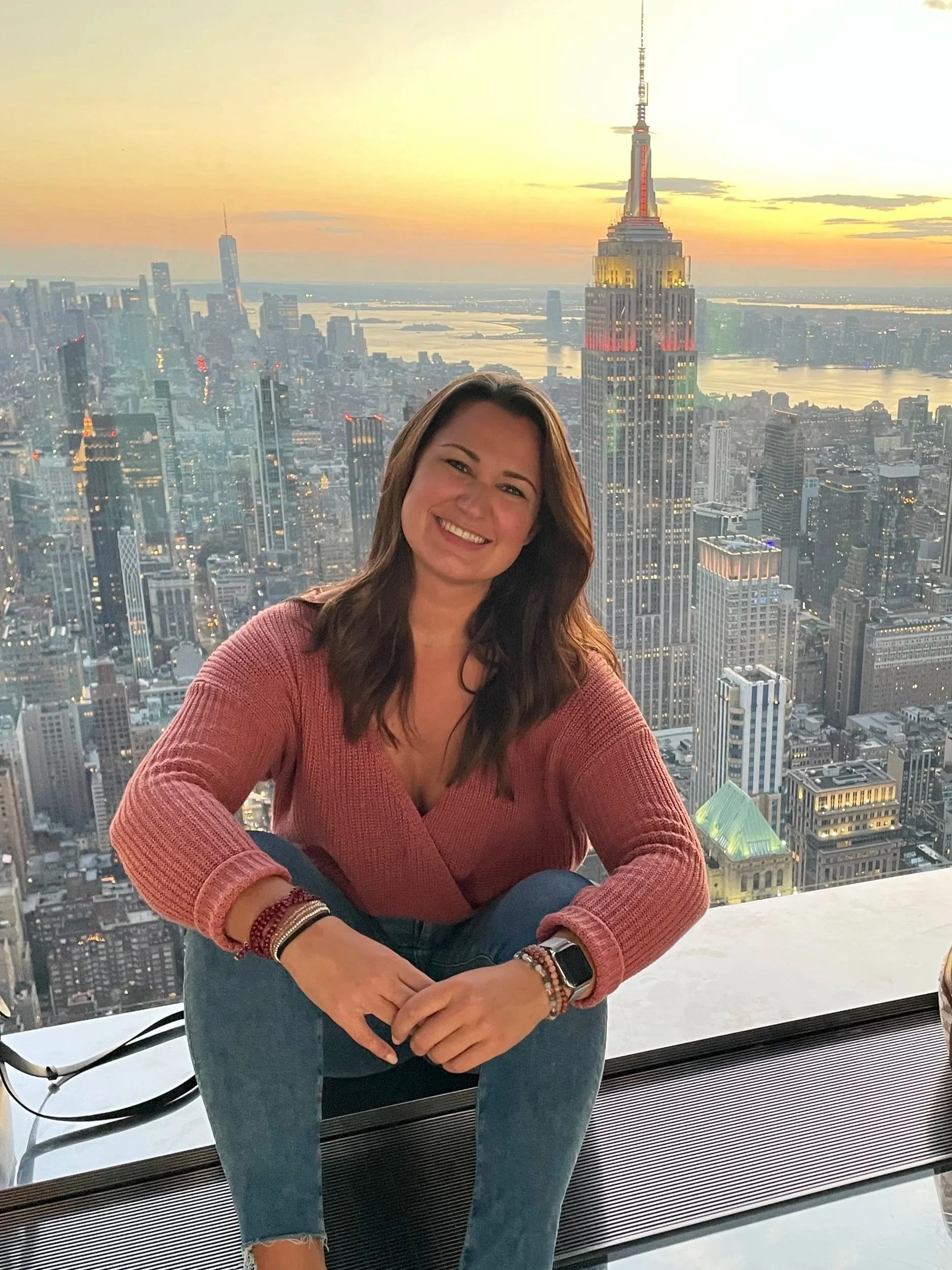 A woman smiling at the camera while sitting on a ledge with the New York City skyline, Empire State Building, and a sunset in the background.