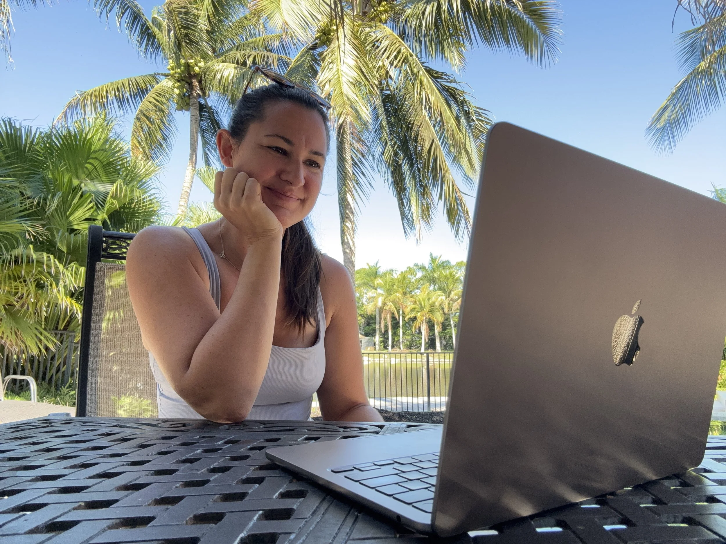 A woman sitting outdoors at a table with a silver MacBook laptop in front of her, surrounded by palm trees and lush greenery, under a clear blue sky.