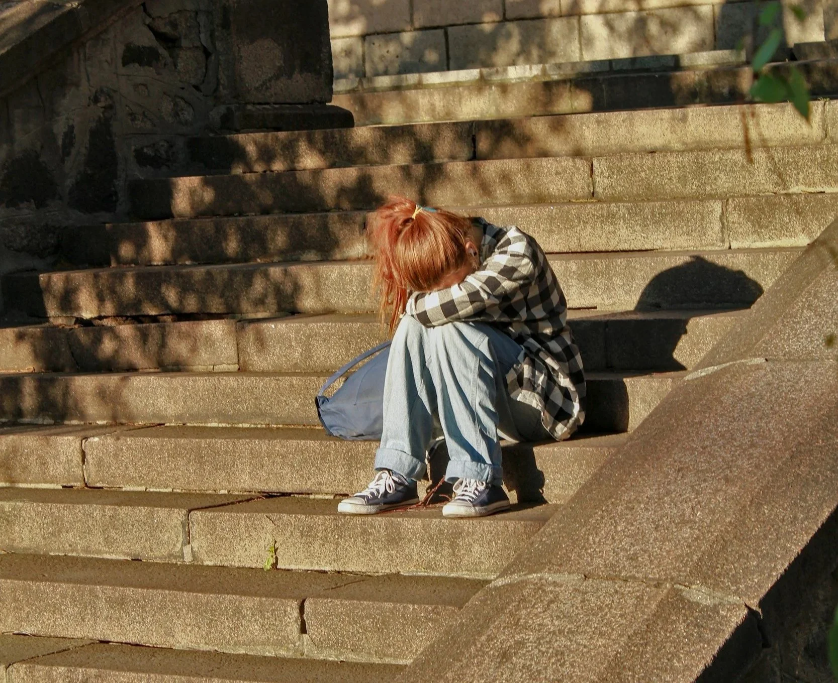 A person with reddish hair sitting on stone steps, wearing a black and white checkered shirt, light blue jeans, and sneakers, with their head down and arms over their knees, holding a bag.