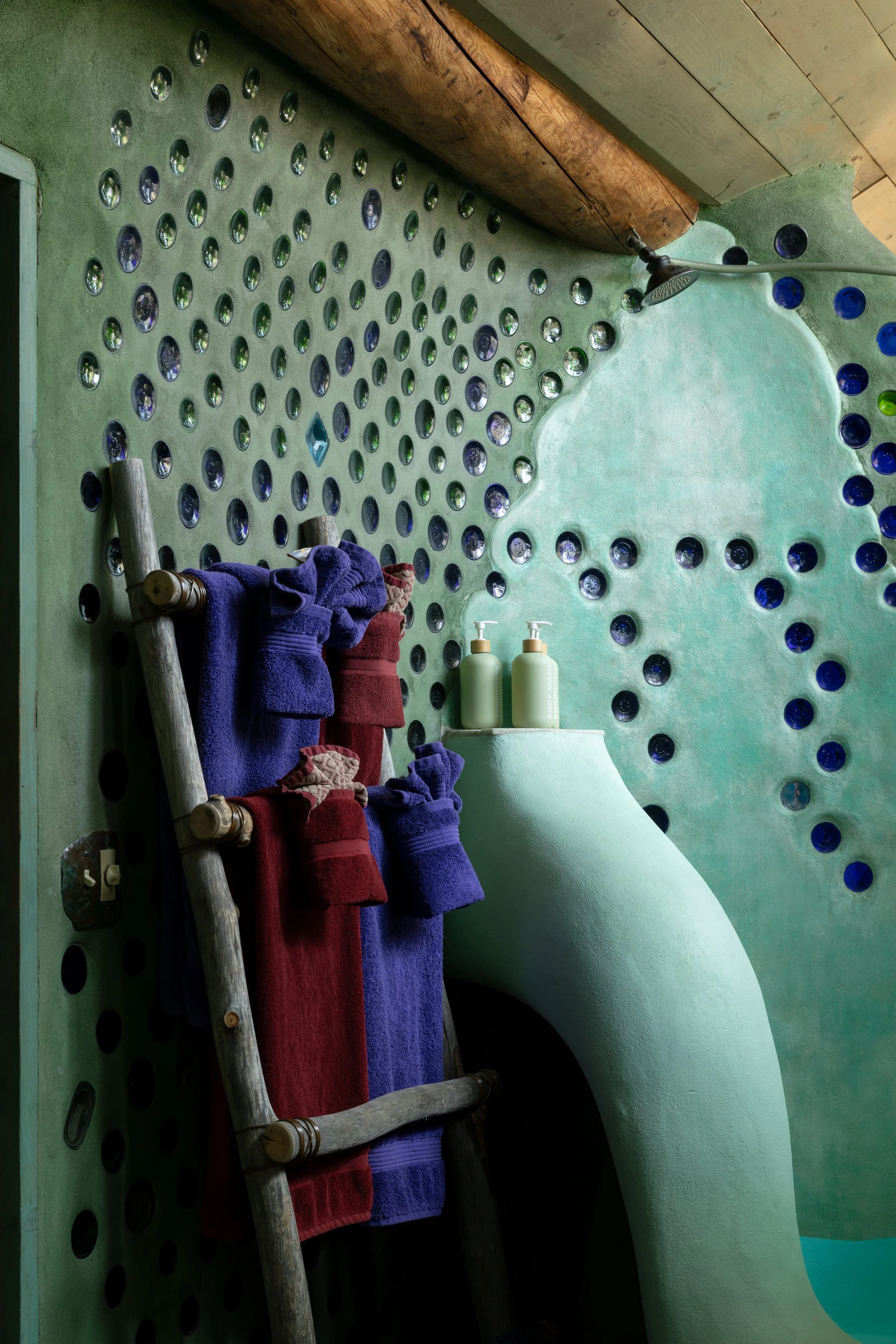 Bathroom with a green wall, decorated with round mirror tiles, a wooden ceiling, and a shower head. A ladder holds purple and red towels, and two soap dispensers sit on a curved structure on the wall.