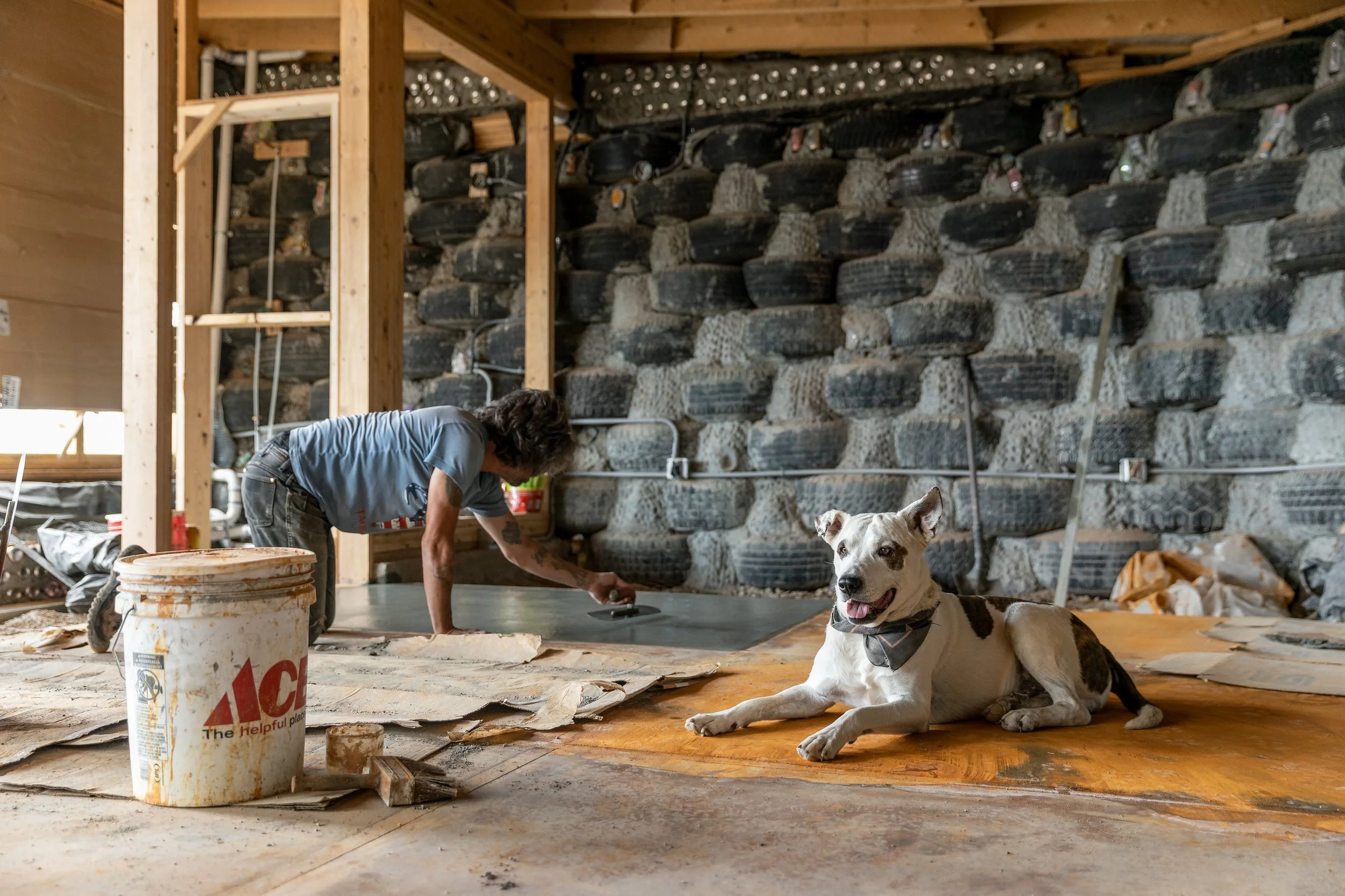A man is working on a concrete floor in a construction site with a dog lying nearby. The dog is white with brown patches and is wearing a collar. The background shows a stone wall and wooden framing.