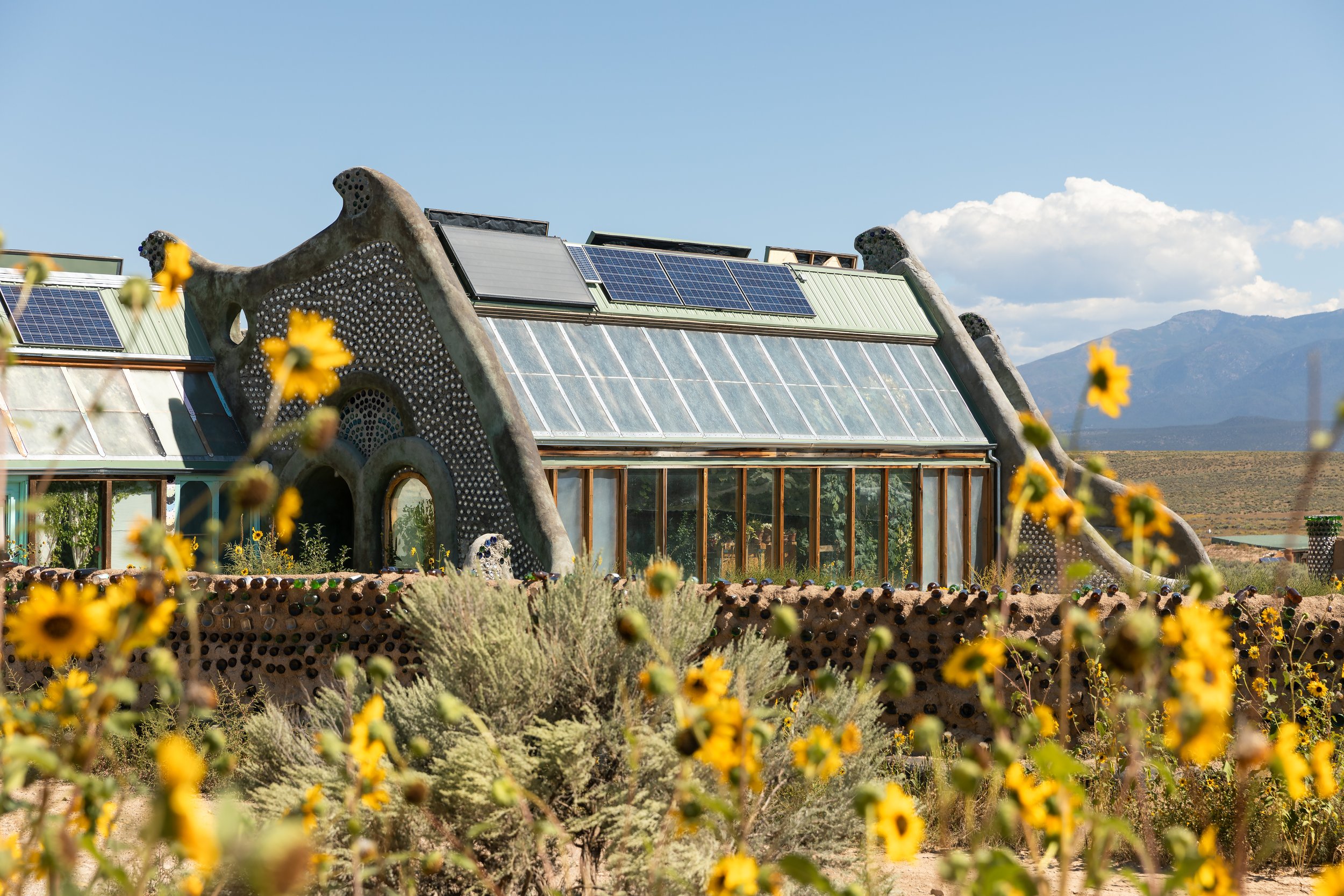 A house with a unique organic design, featuring a curved roof with solar panels and large glass windows, situated in a desert landscape with mountains in the background and yellow flowers in the foreground.