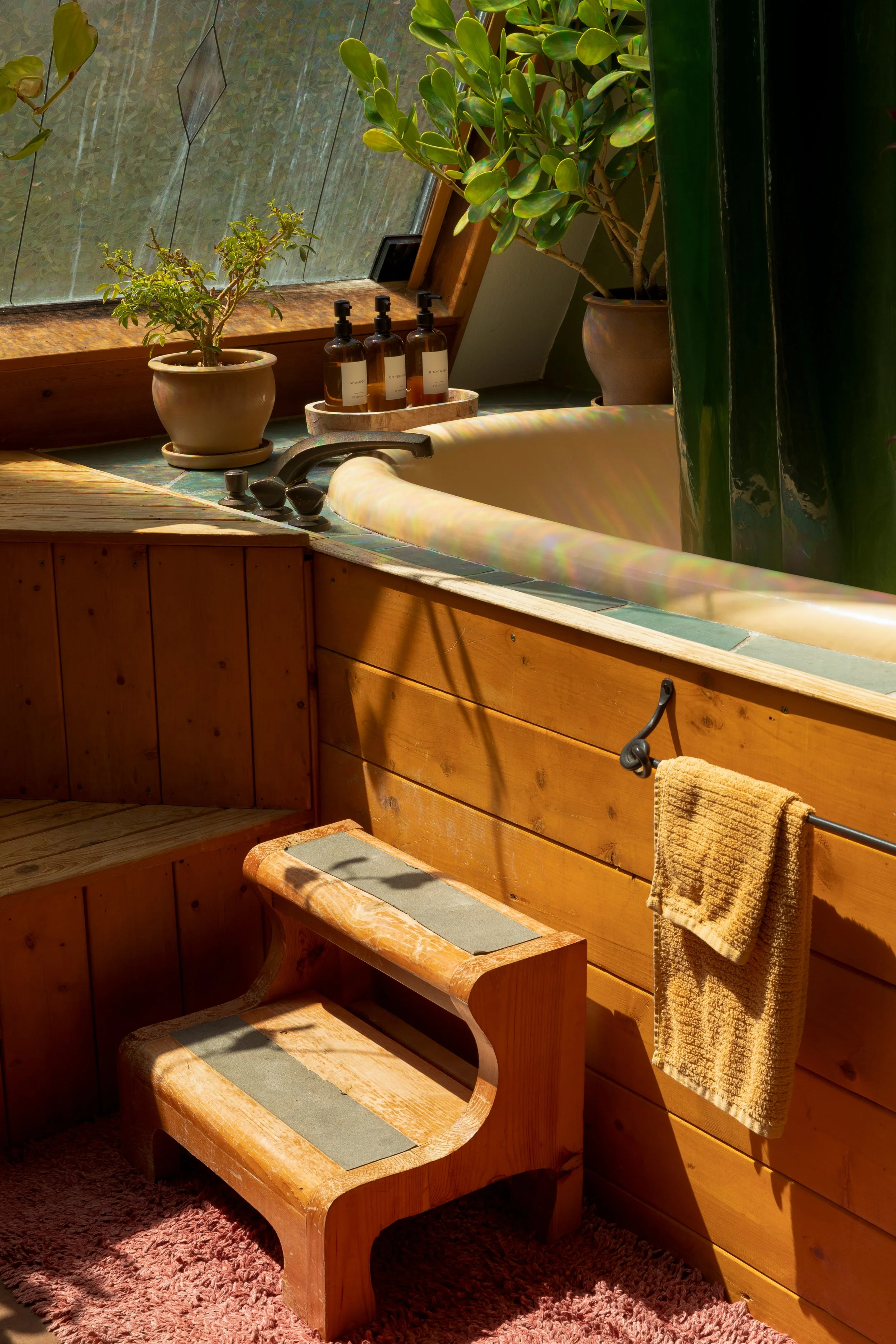 A rustic bathroom with wooden paneling, a corner bathtub, plants on the ledge, and a step stool with a towel hanging on the wall.