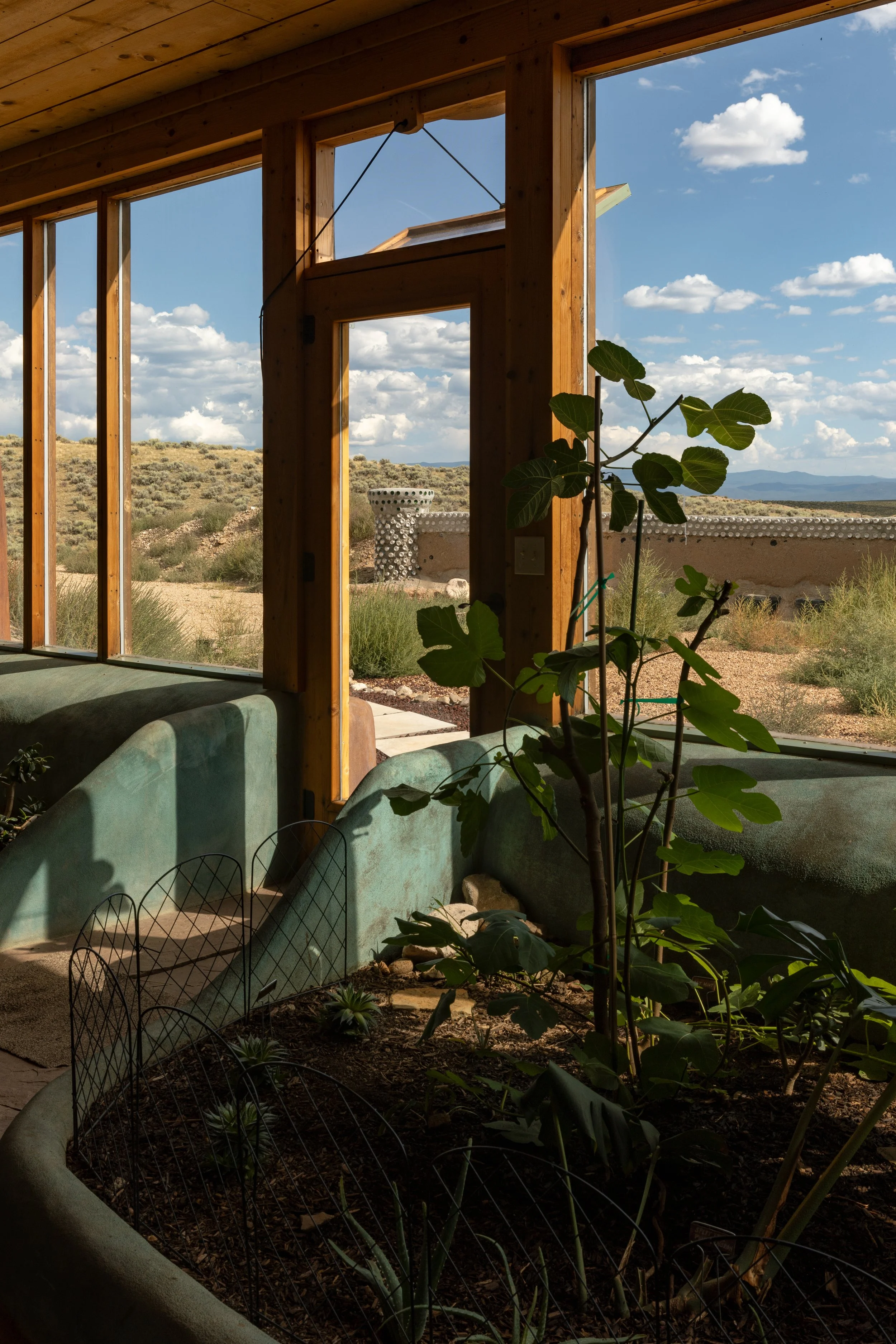 Interior view of a house with large windows showing a desert landscape and blue sky outside. Inside has a potted plant and a built-in bench or wall feature.
