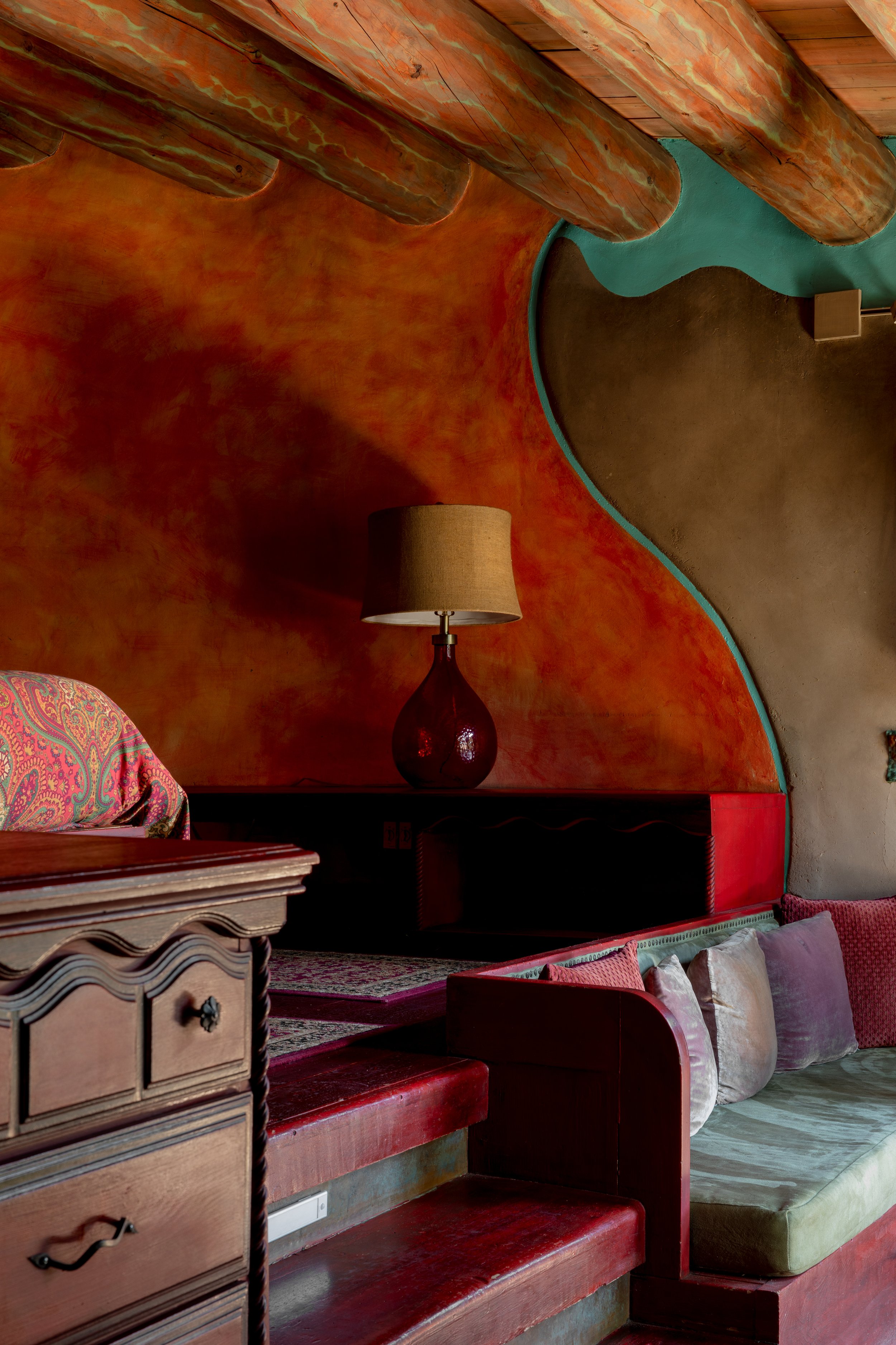 Interior of a room with a colorful, abstract wall, wooden beams on the ceiling, a wooden dresser with ornate drawers, a lamp with a brown shade on a black side table, and a built-in seating area with pillows.